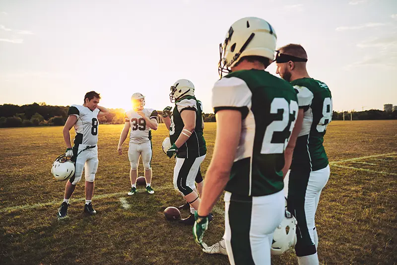 Five young boys standing around after a football player, dressed in football gear